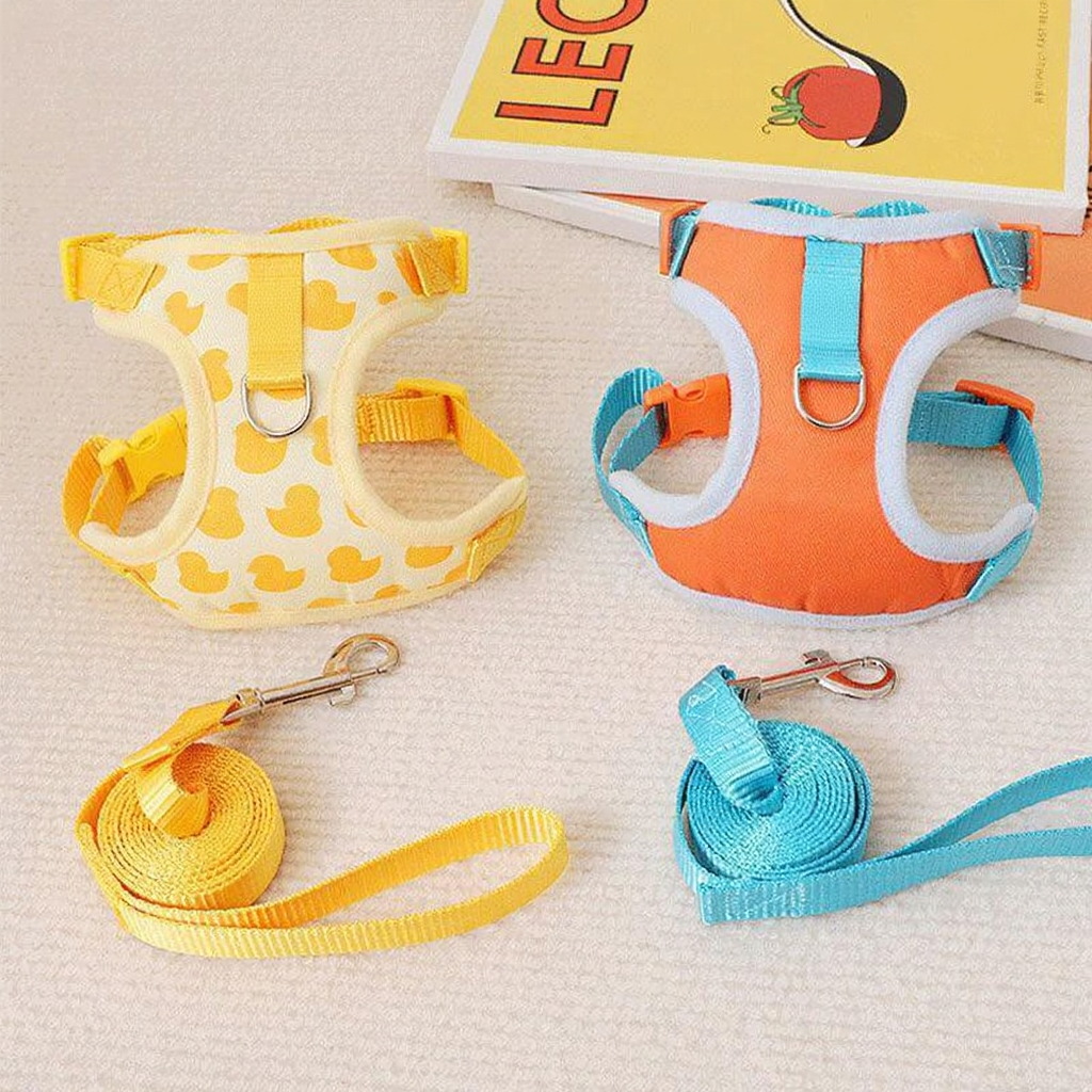 Catvera 3D Animal Escape Proof Harness - Image 11|||PROMPT|||Cleaned image. Original analysis: Found 1 brand logo ('M') on each harness - one on the yellow duck-patterned harness and one on the orange/teal harness, plus a book/product visible in background with branding